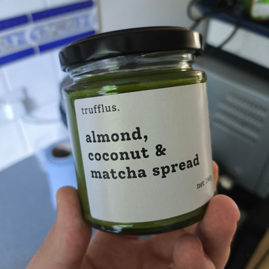 Jar of trufflus almond, coconut & matcha spread held in a hand with a blurred kitchen background.